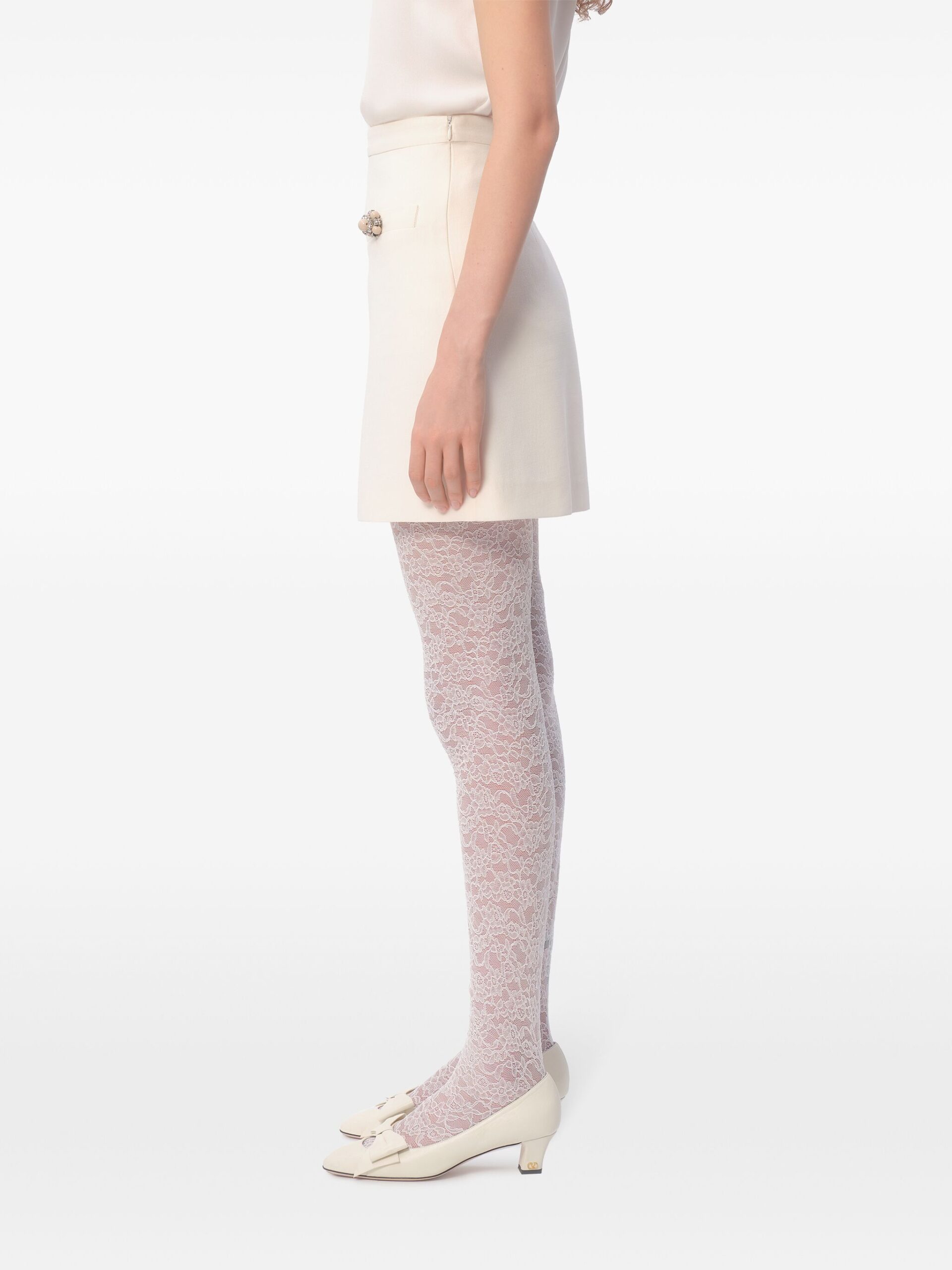 Meio do corpo de mulher com saia creme e meia rendada rosa, usa sapatos brancos com laço, fundo branco.